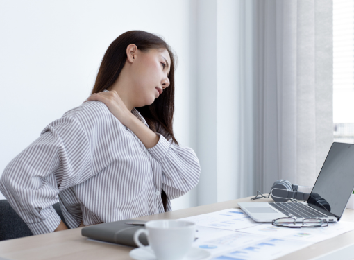 Mulher sentada à mesa de trabalho com expressão de dor, mão no pescoço, ilustrando o impacto da fibromialgia no dia a dia profissional.